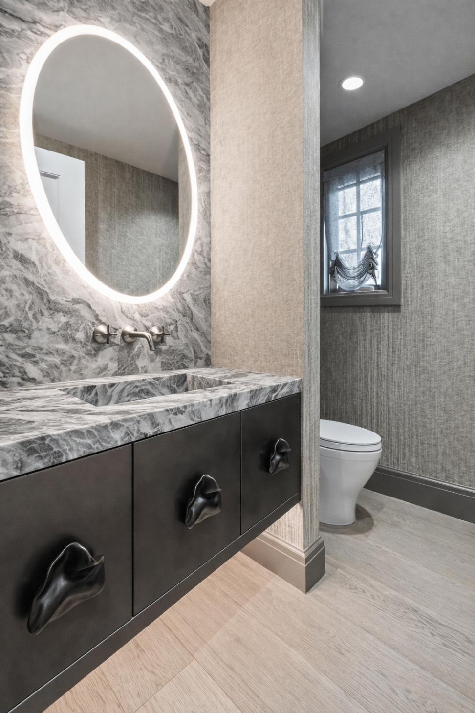 Bathroom with illuminated oval mirror, marble vanity and dark millwork.